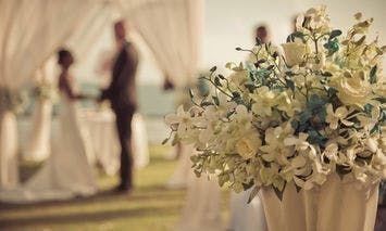 A close-up of a beautifully arranged bouquet of white flowers, likely orchids and roses, in an elegant vase at an outdoor wedding venue.